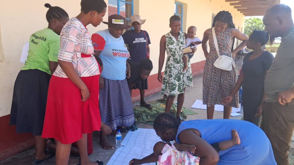 A group of Zimbabwean women stand looking down at sheets of paper in the ground. Two are carrying babies.