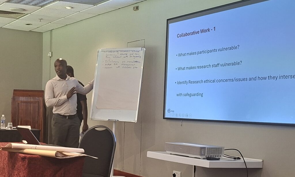 A Zimbabwean man stands next to a flip chart and projection screen, speaking to the room. On the screen are questions for the group around vulnerability and concerns during research