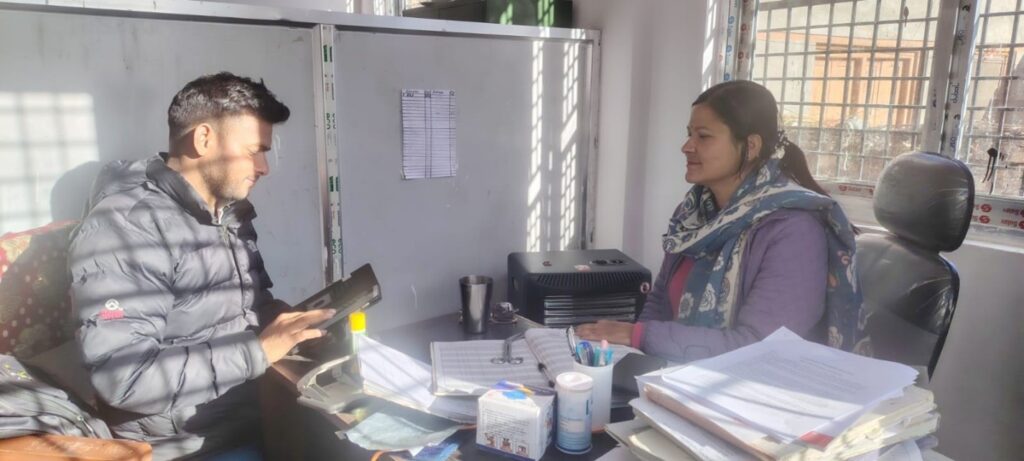 A young Nepali man is a black puffer jacket interviews a Nepali woman behind a desk - she is a doctor