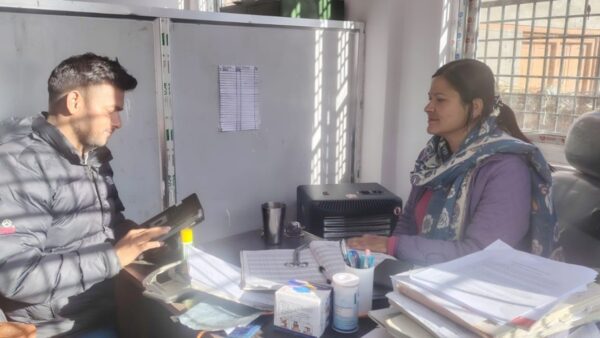 A young Nepali man is a black puffer jacket interviews a Nepali woman behind a desk - she is a doctor
