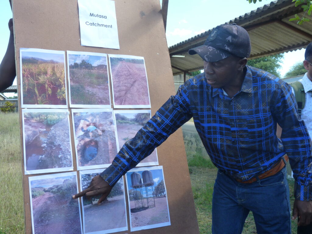 A Zimbabwean man in a chequered shirt and baseball cap points and speaks about to one of 9 photos pinned to a board