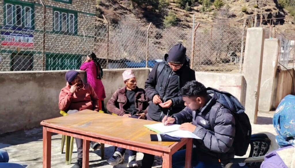 Man in a blac puffer jacket sits outside at a table, writing down the results of interviews with local people, some of whom are gathered nearby