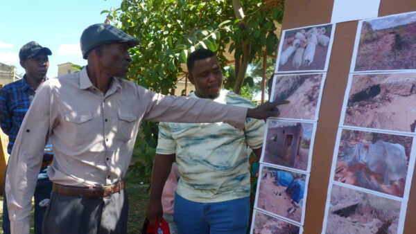 An older Zimbabwean man in a shirt and cap talks to a board of photos he took while others watch on